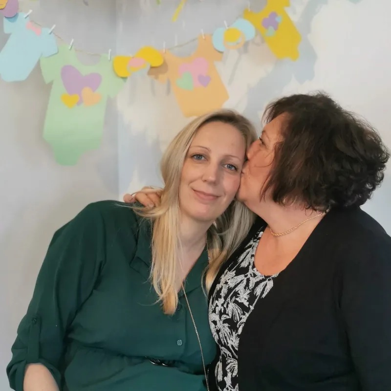 Future maman souriante recevant un baiser sur la joue lors de sa baby shower, sous une guirlande de bodys pastel