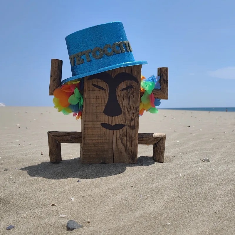 Mascotte totem en bois de Totem, chapeau bleu et colliers de fleurs, plantée sur une plage face à la mer