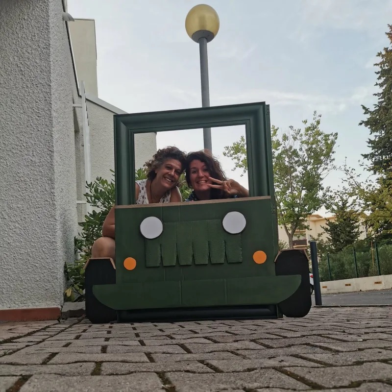 Deux femmes posant dans un photobooth : un cadre en carton en forme de jeep fait main, sourires et signe de la victoire
